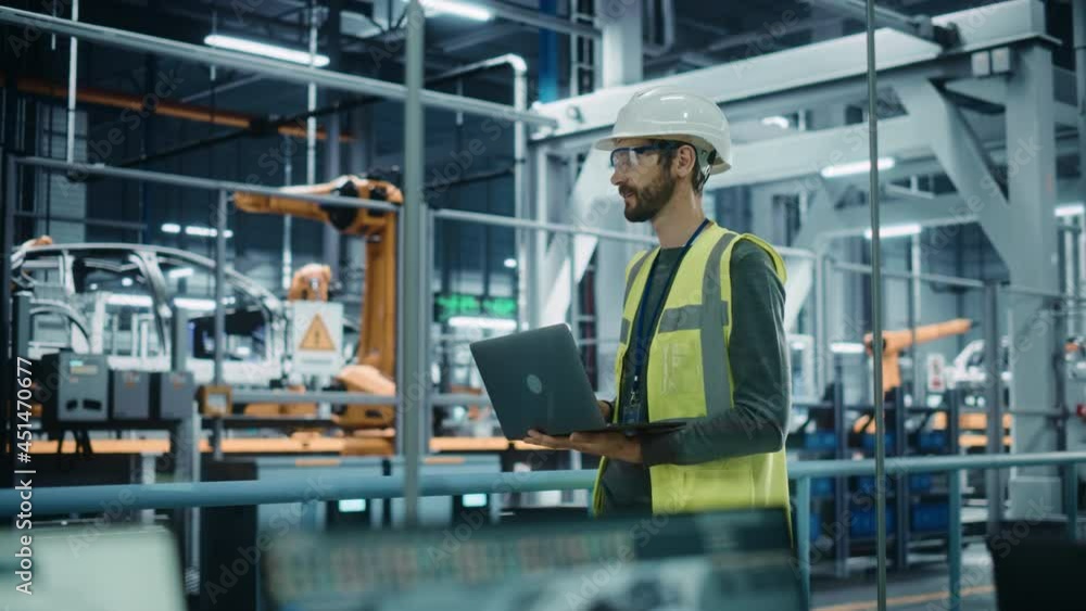 Car Factory: Male Automotive Engineer Wearing Hard Hat, Standing, Using Laptop. Monitoring, Control, Equipment Production. Automated Robot Arm Assembly Line Manufacturing Electric Vehicles. Medium Car Factory: Male Automotive Engineer Wearing Hard Hat, Standing, Using Laptop. Monitoring, Control, Equipment Production. Automated Robot Arm Assembly Line Manufacturing Electric Vehicles. Medium