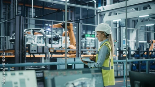 Car Factory: Female Automotive Engineer Wearing Hard Hat, Standing, Using Laptop. Monitoring, Control, Equipment Production. Automated Robot Arm Assembly Line Manufacturing Electric Vehicles. Medium photo