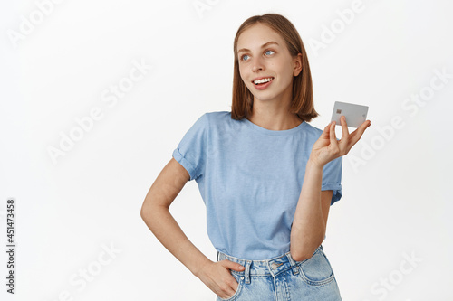 Image of beautiful young woman thinking of going shopping with credit card, looking aside with pleased smile, imaging buying new things, white background
