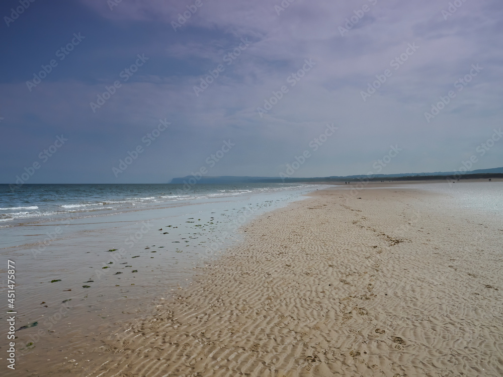 A huge expanse of empty, rippled, sandy beach with footprints leading into the distance and to Hunt Cliff on the horizon, all under a hazy, blue sky.