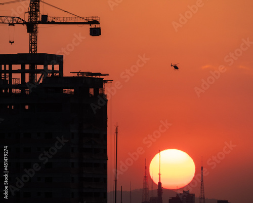 Sol se pondo atrás dos prédios ao fundo e sombra de prédio em construção e primeiro plano com helicopero sobrevoando o céu alaranjado  photo