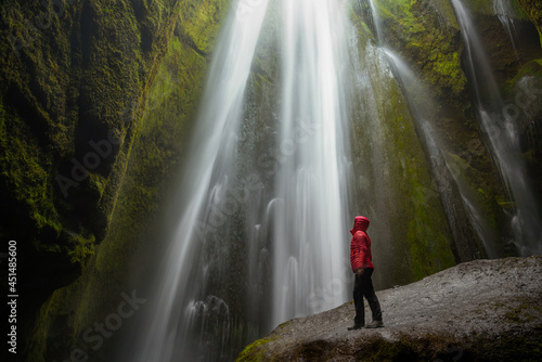 Woman wearing a red jacket standing on a wet boulder at the foot of a majestic waterfall in a narrow canyon. Gljufrabui, Iceland.