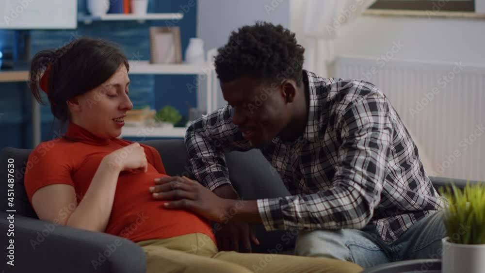 Pregnant interracial couple sitting on living room couch. African american husband touching baby bump and talking to child while caucasian mother relaxing on sofa and laughing Pregnant interracial couple sitting on living room couch. African american husband touching baby bump and talking to child while caucasian mother relaxing on sofa and laughing