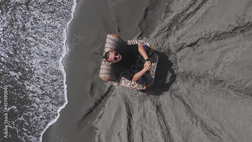 Guy with sunglasses is sitting in old chair in meditative and thoughtful pose, looking up, drone slowly spinning around, top view. Relaxing recreation after hard work or before important life changes. photo