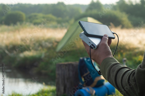 Fototapeta Naklejka Na Ścianę i Meble -  Man holds a smartphone in his hands and charges it with a power bank against the backdrop of a tourist tent in nature. Portable travel charger.