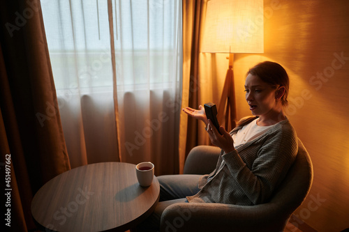 A young woman is sitting in a chair with a phone in her hand. He looks at the phone in surprise. There is a cup of coffee on the table. Hotel, rest, conversation, emotions.