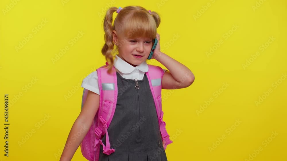 Dissatisfied nervous schoolgirl with backpack making a call with mobile phone to mother, discussing very irritated aggressive over yellow studio wall background. Family quarrel. Back to school concept Dissatisfied nervous schoolgirl with backpack making a call with mobile phone to mother, discussing very irritated aggressive over yellow studio wall background. Family quarrel. Back to school concept
