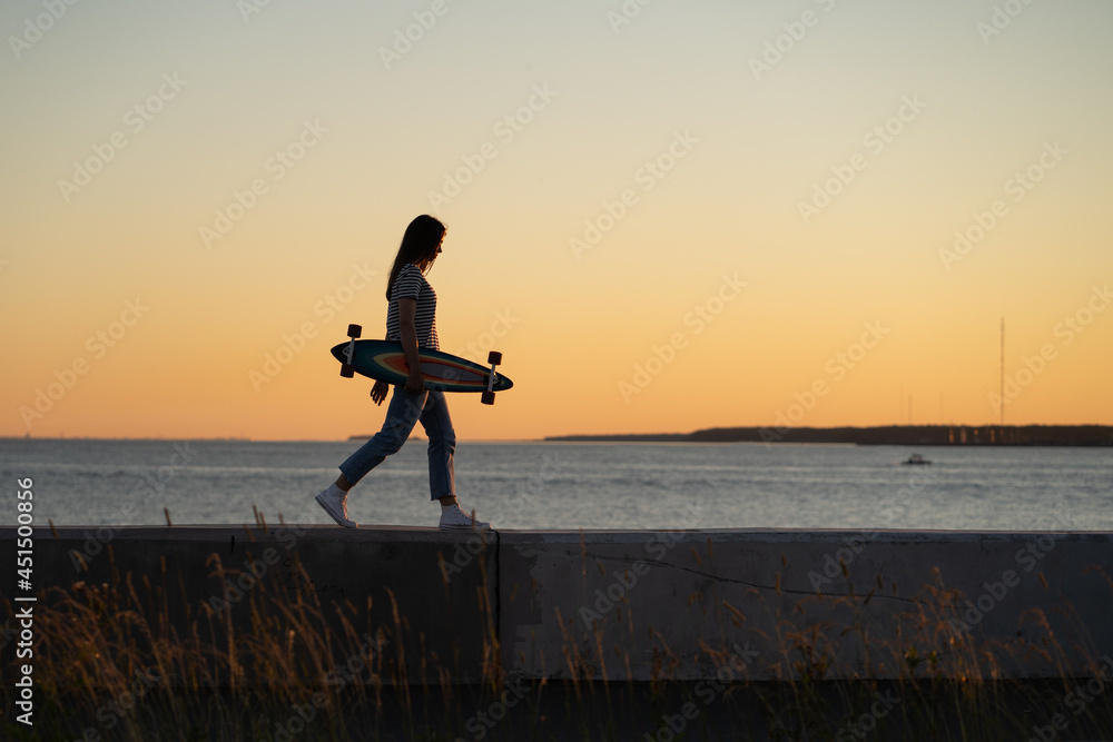 Lifestyle and freedom concept: young girl with longboard walking over sunset view. Silhouette of trendy teen or student female skater with skateboard at riverside pavement. Woman walk along river
