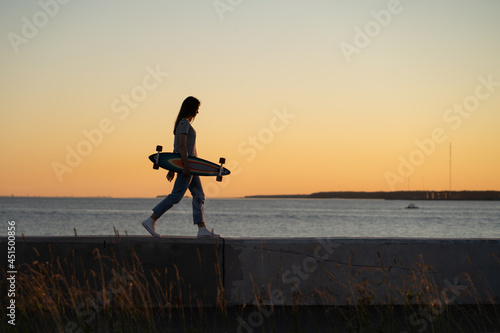Lifestyle and freedom concept  young girl with longboard walking over sunset view. Silhouette of trendy teen or student female skater with skateboard at riverside pavement. Woman walk along river