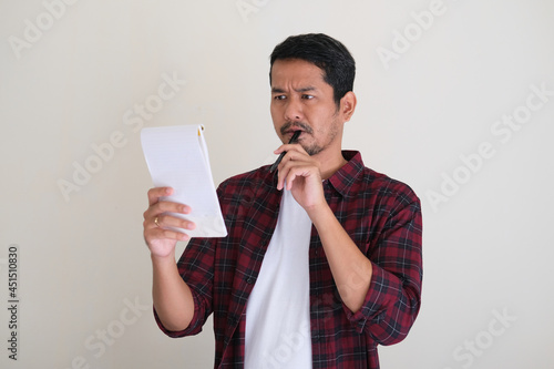 Asian man bite a pen while reading notes showing confused expression photo