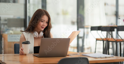 Smiling girl student wear wireless headphone study online with teacher, happy young asian woman learn language listen lecture watch webinar write notes look at laptop sit in cafe, distant education.