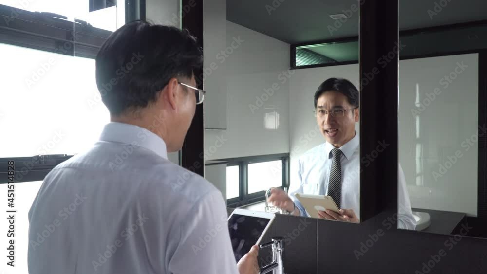 Asian businessman standing in front of mirror practicing speech with looking text script on digital tablet with smiling face. Male speaker training speaking skill for presentation in conference room Asian businessman standing in front of mirror practicing speech with looking text script on digital tablet with smiling face. Male speaker training speaking skill for presentation in conference room