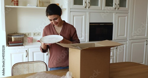 Indian woman standing in kitchen unpack parcel box with ordered fragile dishware crockery items wrapped in bubble wrap smiles feels satisfied by safe transport and e-commerce retail services concept