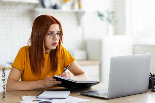 Online education. Focused young red-haired woman looks at the laptop screen and takes notes in a notebook. Remote learning, work or training online concept