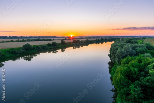 Fototapeta Naklejka Na Ścianę i Meble -  Scenic view at beautiful summer river sunset with reflection on water with green bushes, grass, golden sun rays, calm water ,deep cloudy sky and glow on a background, spring , evening landscape