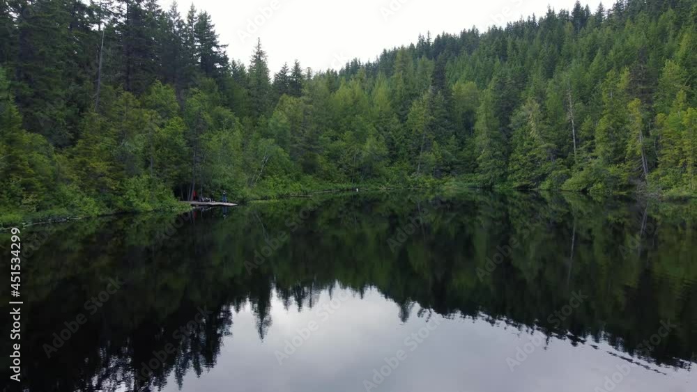 Flight over mirror lake surrounded by green forest in Canada on an overcast day Flight over mirror lake surrounded by green forest in Canada on an overcast day