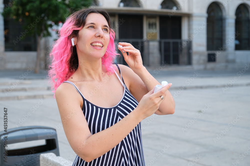 Young caucasian woman with pink hair putting wireless earbuds in street.