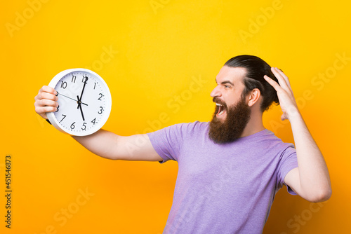 Photo of frustrated bearded man holding clock, screaming and geasturing over yellow background