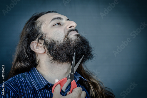 portrait of a young man with a beard and long hair who, with a scissor in his hand, wants to change his look and wants to cut his beard, concept to illustrate the desire to change photo