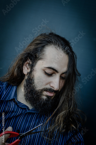 portrait of a young man with a beard and long hair who, with a scissor in his hand, hesitates to cut his hair, concept to illustrate the desire to change photo