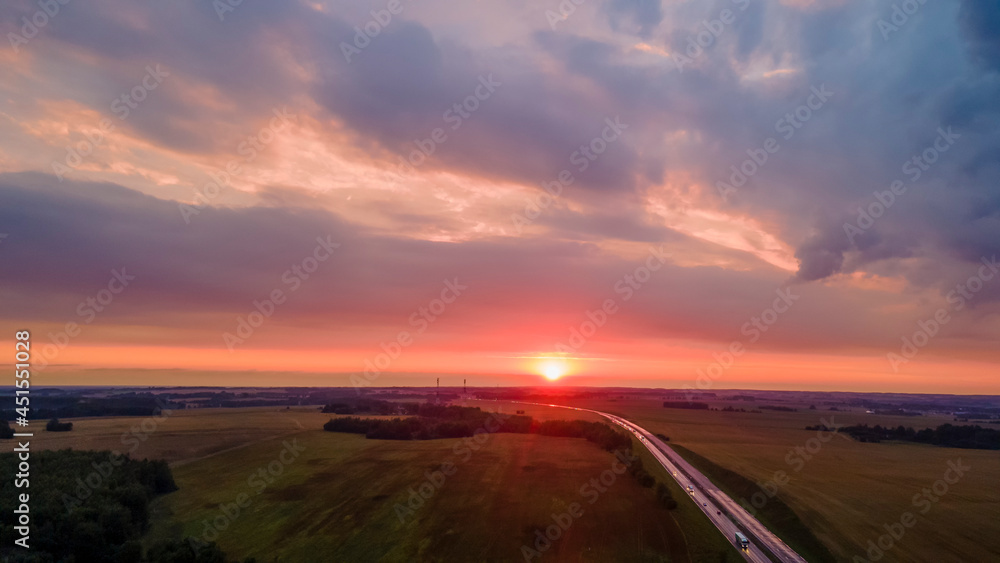 Aerial view of highway on red sunset. Landscape with road near countryside fields. Beautiful winding road leading through rural countryside with evening sunlight. Dramatic sky background.