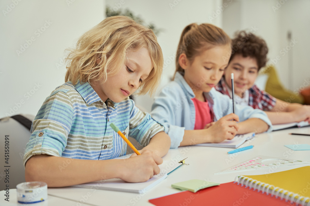 Quality education. Diligent schoolboy making notes during lesson. Kids studying, sitting together at the table in elementary school classroom Quality education. Diligent schoolboy making notes during lesson. Kids studying, sitting together at the table in elementary school classroom