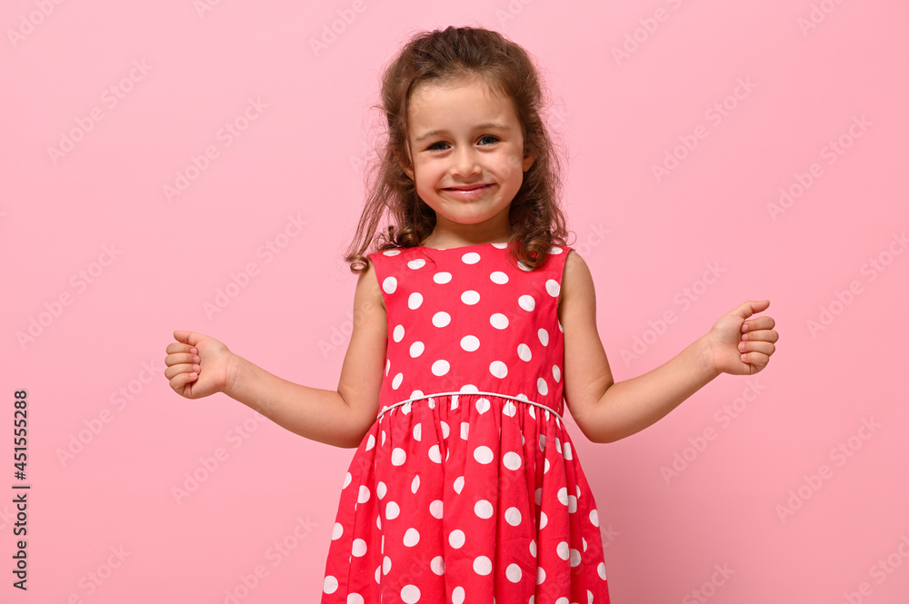 Face portrait of a beautiful child girl in pink polka dots dress, cute smiling looking at camera, posing with thumbs up, over pink background with copy space Face portrait of a beautiful child girl in pink polka dots dress, cute smiling looking at camera, posing with thumbs up, over pink background with copy space