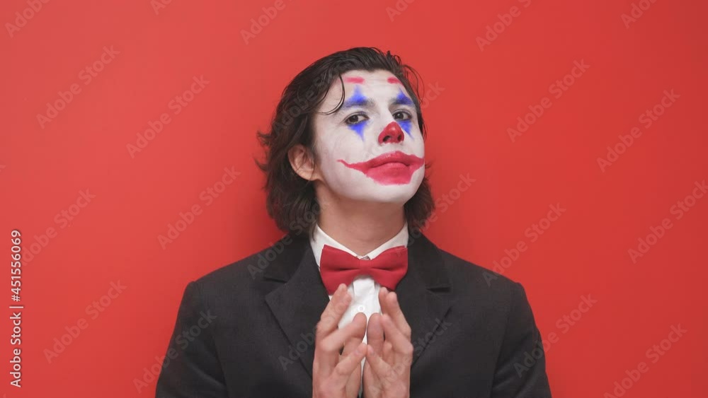 A clown with colorful makeup poses on an isolated red background, a joyful magician or a mysterious man in a black suit, talks, smiles A clown with colorful makeup poses on an isolated red background, a joyful magician or a mysterious man in a black suit, talks, smiles