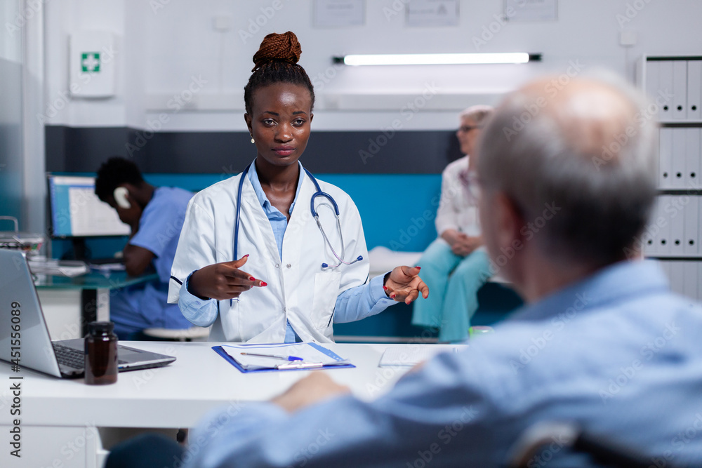 Doctor of african american ethnicity talking to elder man about disease and healing treatment at healthcare clinic in office. Black medic discussing with old sick patient while sitting at desk Doctor of african american ethnicity talking to elder man about disease and healing treatment at healthcare clinic in office. Black medic discussing with old sick patient while sitting at desk