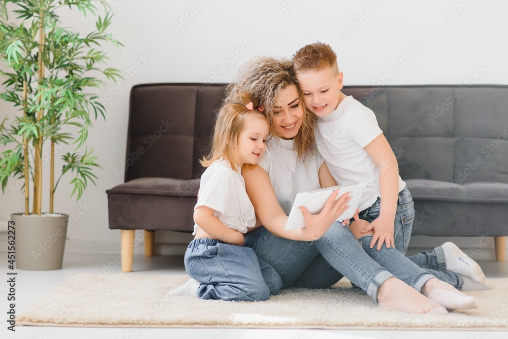 Beautiful young mother, their cute little daughter and son are using a tablet and smiling, sitting on the floor at home