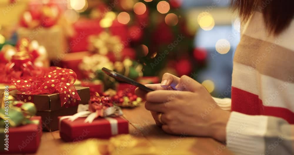 Unrecognizable woman in thick winter clothes sitting and desk with a pile of gift boxes and typing messages on smartphone screen with Christmas tree and bokeh lights in blur background. Unrecognizable woman in thick winter clothes sitting and desk with a pile of gift boxes and typing messages on smartphone screen with Christmas tree and bokeh lights in blur background.