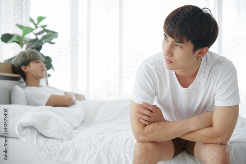 Lgbt Asian gay couple quarreling and irritated in bedroom. Stressed young man sitting at end of bed with arms crossed and boyfriend made sullen face and lay in bed. Homosexual in problem relationship