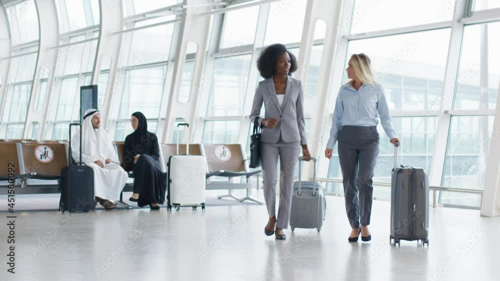Caucasian attractive businesswoman in business style and her multiracial colleague walking through the corridor of the airport with suitcases, speaking and waiting for the plain Caucasian attractive businesswoman in business style and her multiracial colleague walking through the corridor of the airport with suitcases, speaking and waiting for the plain