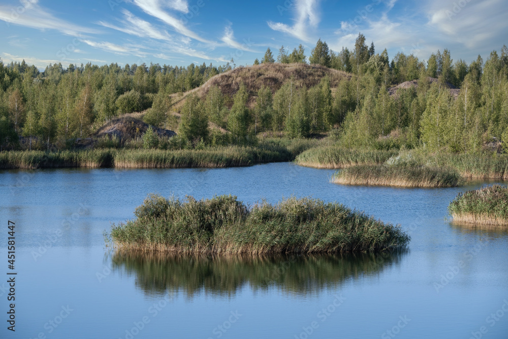 A lake with a plane surface and reflections against the blue sky and white clouds. Originated from an old quarry flooded, groups of islands overgrown with reeds. Beauty in nature, landscapes, scenic.