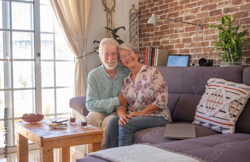 Smiling senior couple at home sitting on sofa looking at camera. Brick wall on background