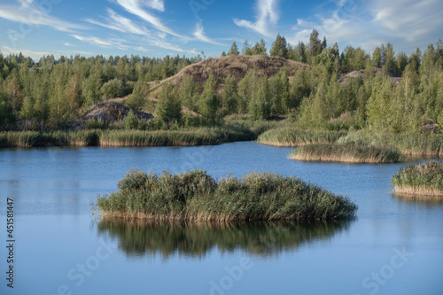 Fototapeta Naklejka Na Ścianę i Meble -  A lake with a plane surface and reflections against the blue sky and white clouds. Originated from an old quarry flooded, groups of islands overgrown with reeds. Beauty in nature, landscapes, scenic.