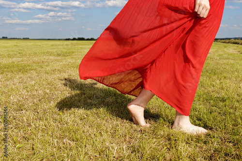Woman in red skirt is walking barefoot on grass summer meadow. Cropped person view.
