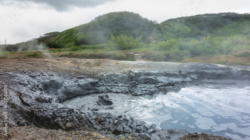 In the valley of hot springs, the mud boiler is filled with a gray liquid. There are bubbles and steam above the surface. Clay edges. Green mountains against a cloudy sky. Kamchatka