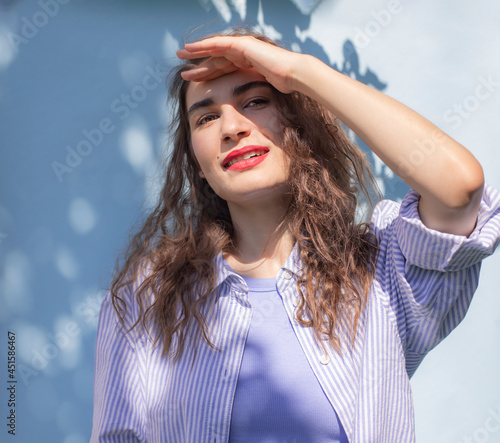 Happy young beautiful brunette in blue oversize shirt with curly hair looking into the camera and hiding her face from the harsh sun with her hand. Creative modern outdoor portrait  with tree shadows
