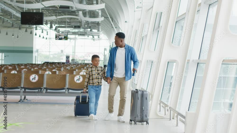 Multiracial nice and happy father and son walking the airport corridor with suitcases on wheels and smiling while going to the vacation Multiracial nice and happy father and son walking the airport corridor with suitcases on wheels and smiling while going to the vacation