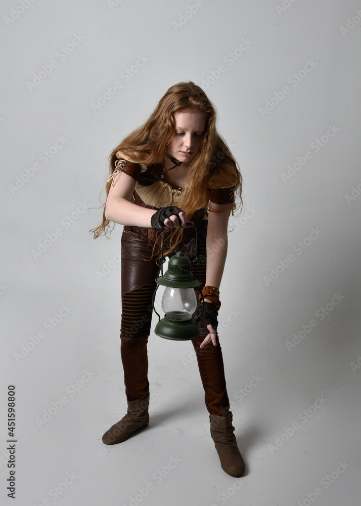 Full length close up portrait of beautiful young woman with long red hair, wearing steampunk inspired costume holding lantern isolated on studio background. Full length close up portrait of beautiful young woman with long red hair, wearing steampunk inspired costume holding lantern isolated on studio background.