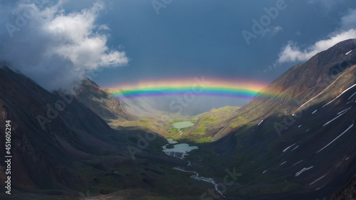 Full rainbow over a mountain valley. Atmospheric alpine landscape with snowy mountains with rainbow in rainy and sunny weather.