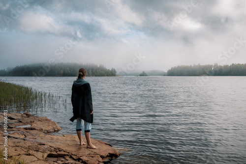 A girl with long hair in a blue jacket stands barefoot on a large stone on the shore of the lake and looks at the beautiful islands in the fog.