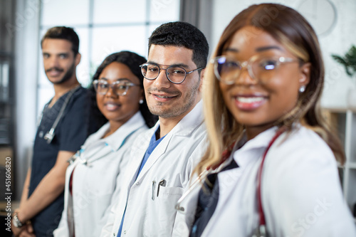 Close up portrait of happy cheerful multiethnic medical team standing in a row in the hospital. Focus on handsome male Arabic Indian physician smiling at camera