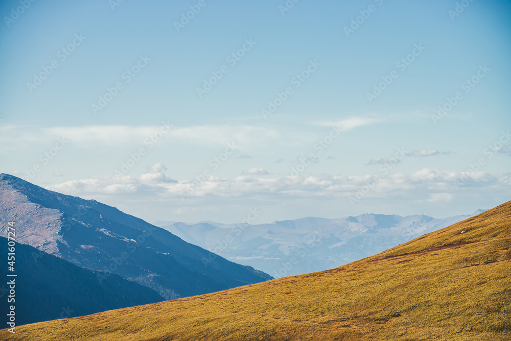Minimalist autumn landscape with diagonal of sunlit orange mountainside on background of mountains silhouettes on horizon. Minimal mountain scenery with slope of hill in golden sunlight in autumn time
