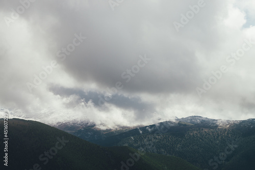 Atmospheric autumn landscape with forest mountains in snow in low clouds in overcast weather. Awesome scenery with gray rain clouds above snowy hills and mountains in autumn colors. Grainy clouds.