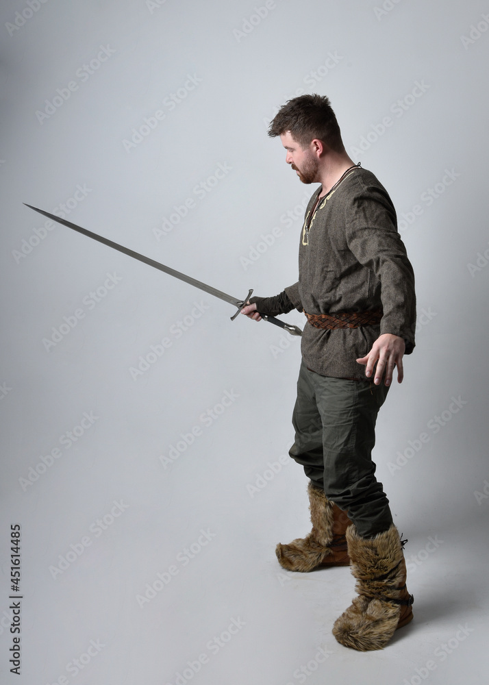 Full length portrait of young handsome man wearing medieval Celtic adventurer costume. Standing pose holding a sword, isolated on studio background. Full length portrait of young handsome man wearing medieval Celtic adventurer costume. Standing pose holding a sword, isolated on studio background.