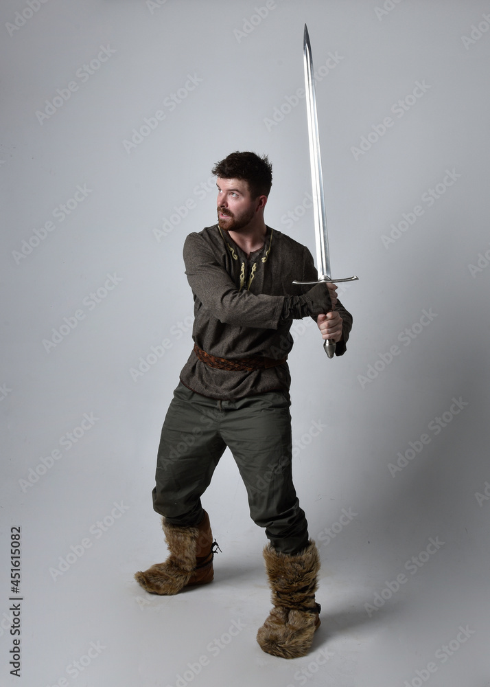 Full length portrait of young handsome man wearing medieval Celtic adventurer costume. Standing pose holding a sword, isolated on studio background. Full length portrait of young handsome man wearing medieval Celtic adventurer costume. Standing pose holding a sword, isolated on studio background.