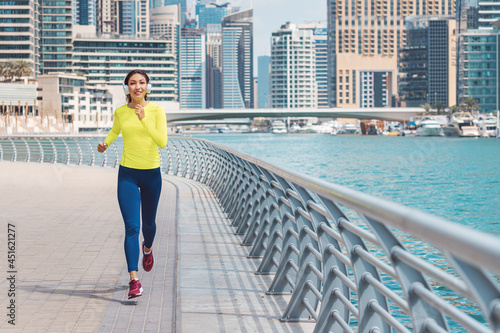 Happy woman wearing headphones and sunglasses trains and does running and fitness on the sidewalk of the embankment in the Dubai Marina area © EdNurg