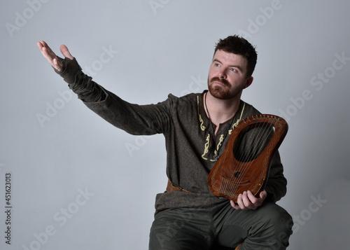 Full length  portrait of  young handsome man  wearing  medieval Celtic adventurer costume playing a small musical harp, isolated on studio background. photo